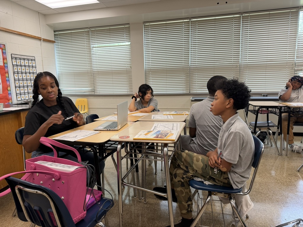 Several people seated around desks in a classroom. A woman with a pen and a laptop.