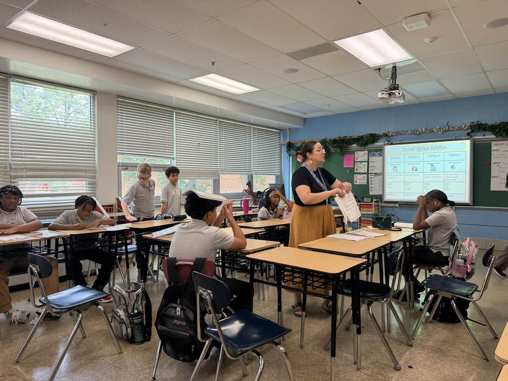 In a classroom, a teacher stands at a desk, addressing a group of students seated at their desks.
