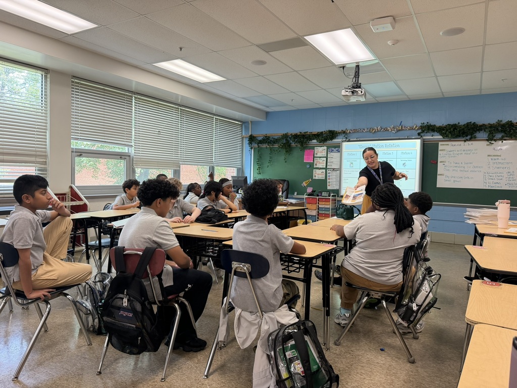 A classroom with a teacher standing near a projector screen. Students sit in rows at tables, some wearing backpacks.