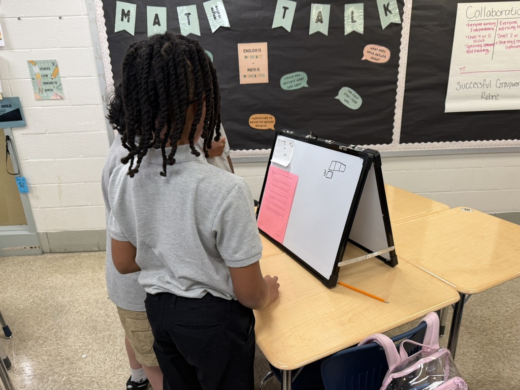 A student stands before a whiteboard with a tablet on it, possibly presenting. Behind them, a bulletin board with "Math Talk" written on it.