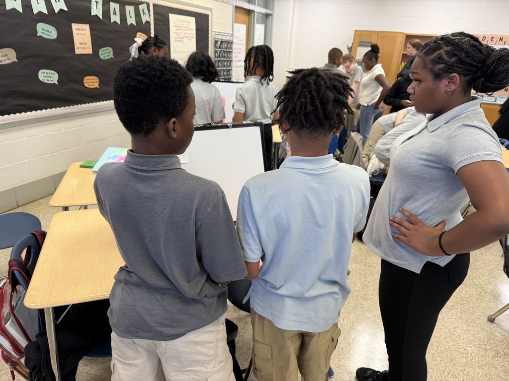Classroom with children and teacher. Two boys are discussing something with teacher. Various tables, chairs, and bulletin board in background.