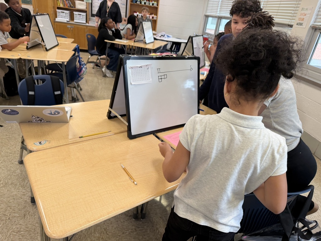 A classroom with children sitting at tables. A girl in a white shirt stands near a whiteboard, writing with a marker.