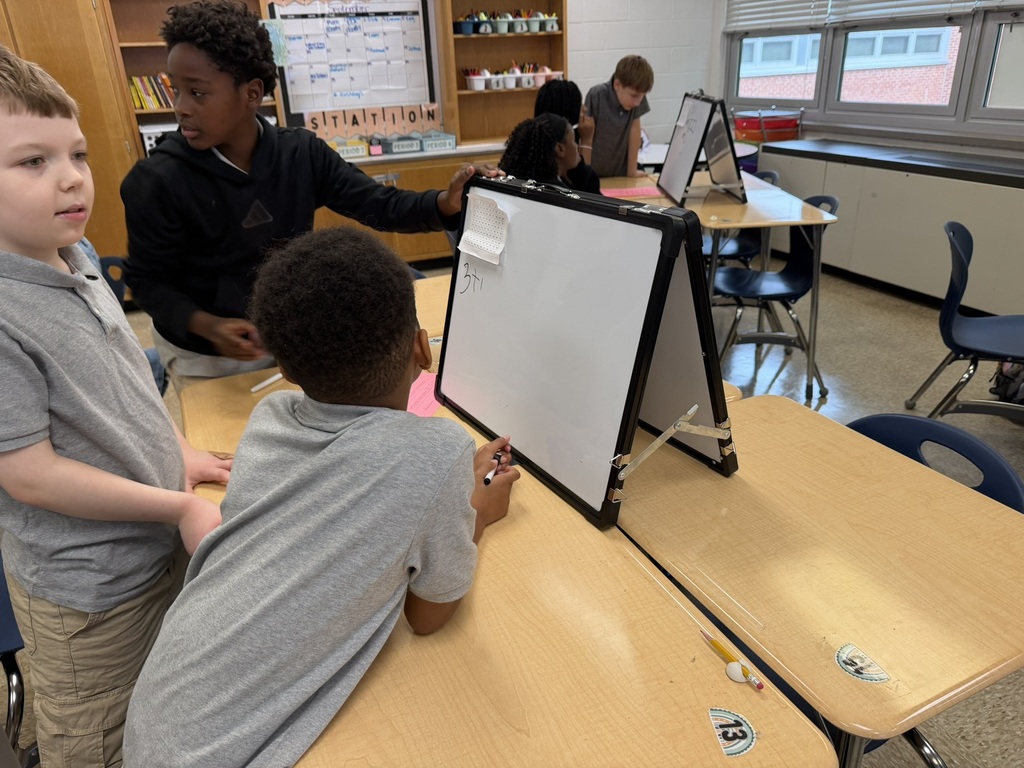 Children stand around a desk with a whiteboard. One child writes on the board.