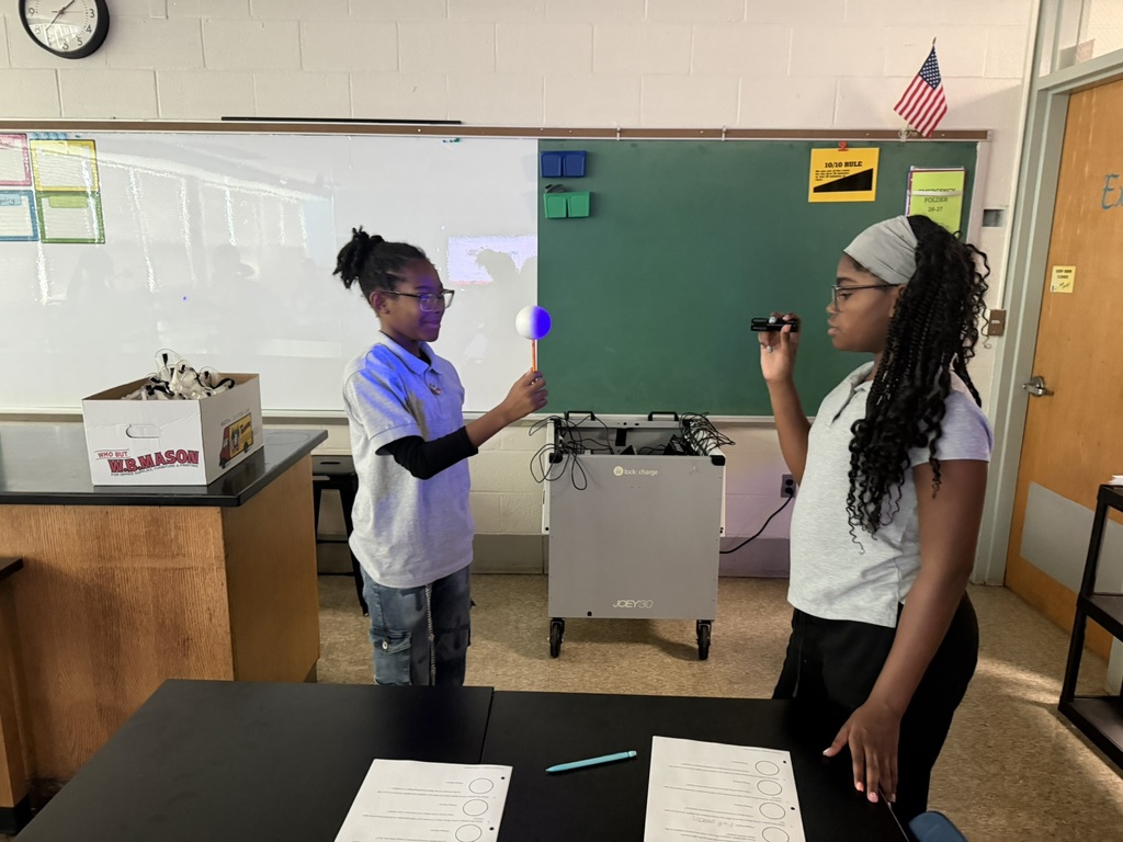Two students hold flashlights in a classroom. One student is in front of a table, and the other is in front of a board.