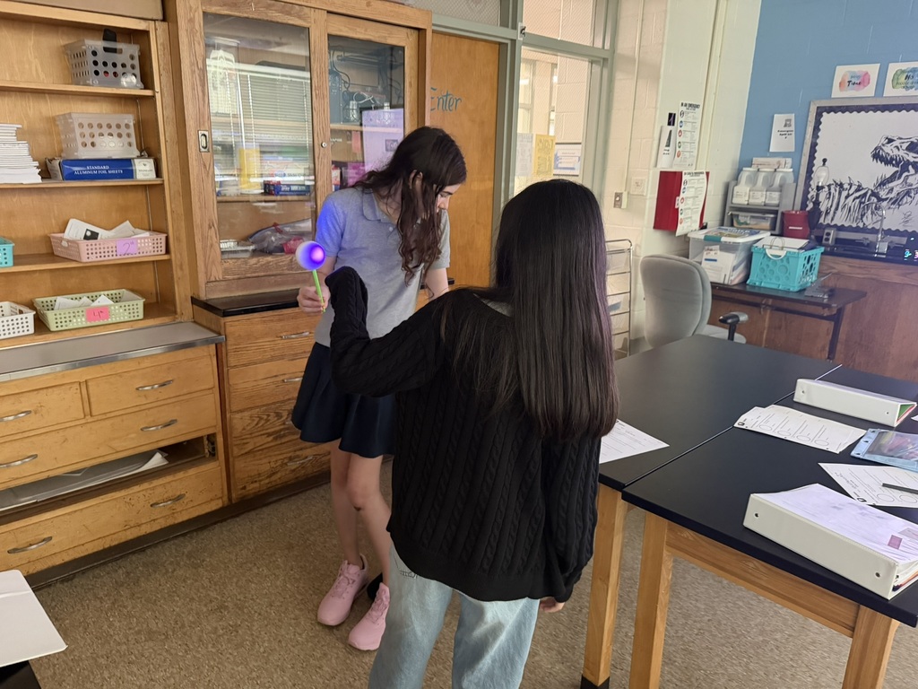 Two individuals in a classroom with one holding a purple light. Shelves, a desk, and papers are in the room.