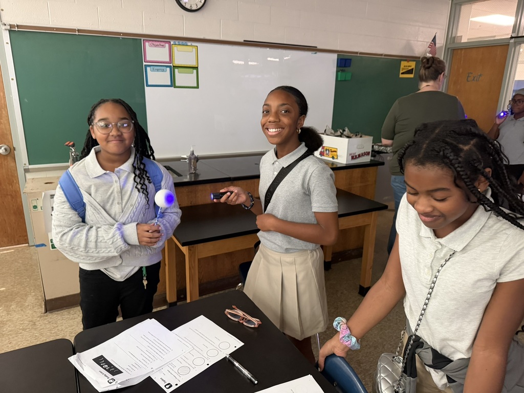 Three students stand in a classroom, two holding objects, one at a desk, another next to a whiteboard.