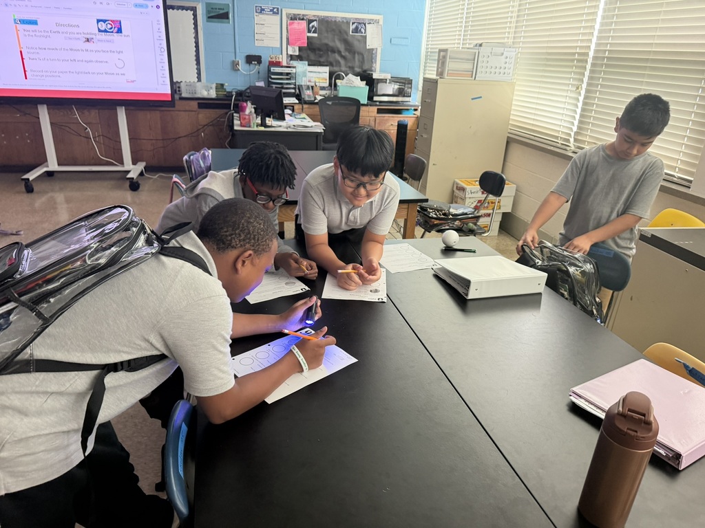 Four young students in a classroom are engaged in activities around a table. One student is using a tablet.