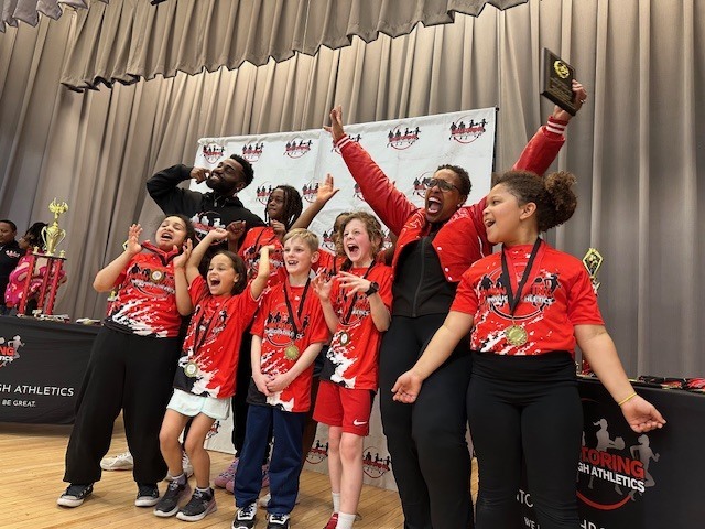 A team of children in red tops, shorts, and sneakers with coaches celebrate in front of a banner.