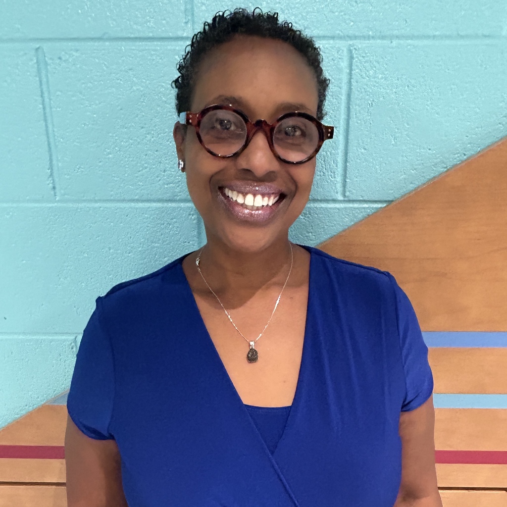 A smiling woman with glasses and a necklace stands in front of a blue wall.
