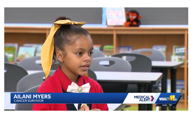 A girl with a yellow headband sits in a classroom, facing the camera. Behind her are school desks and shelves.