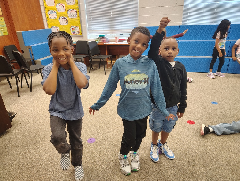 Three children in a classroom, two standing and smiling while the third lies on the floor.