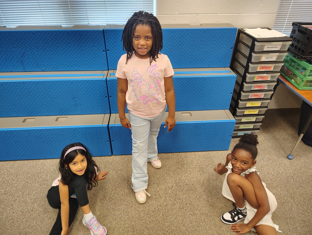 Three young girls are posing in front of blue bleachers with a table and storage boxes behind them.