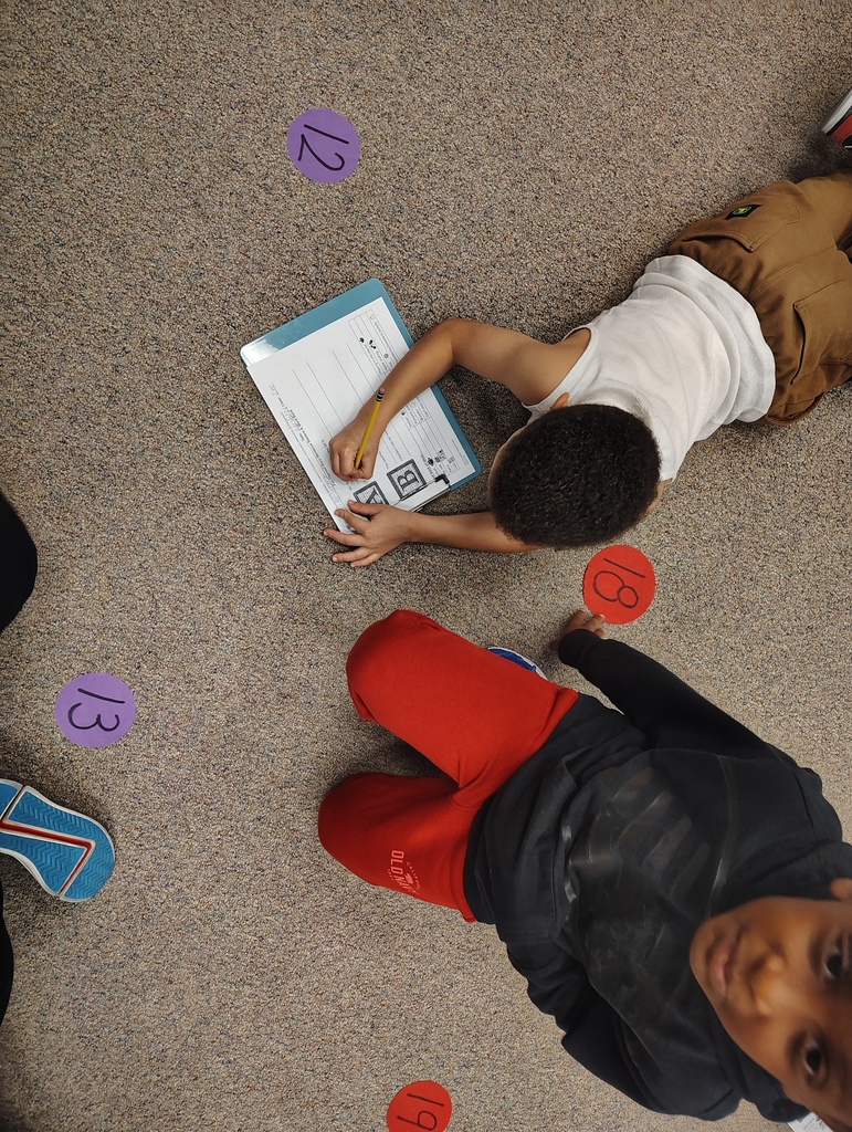 Two children on floor with numbered dots, one lying down writing in a book, other lying down watching.