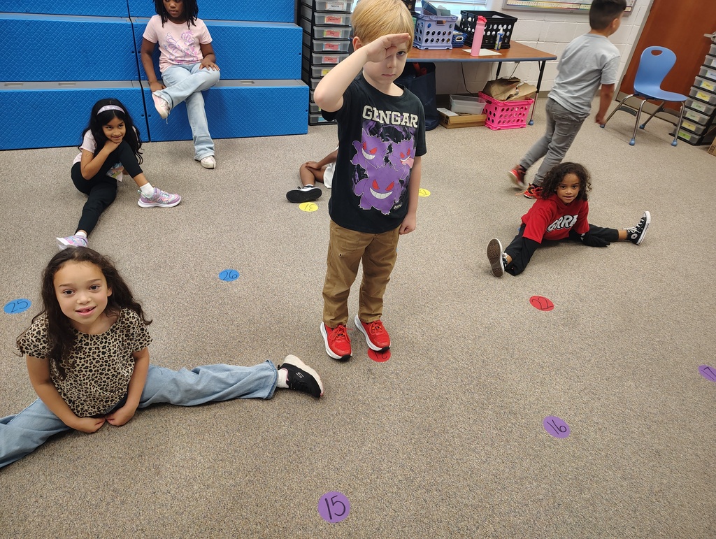Kids play in a carpeted room with colored circles on the floor. Some sit, others stand. A boy salutes.