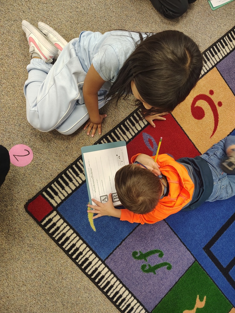 Two children, one wearing orange, lie on a colorful floor mat, studying a book. A musical note is visible on the mat.