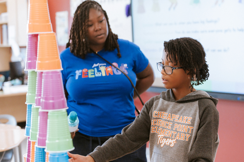 AACPS Mentoring Conference 2026: A young person in a hoodie builds a tower of colored cups while a teacher stands behind.