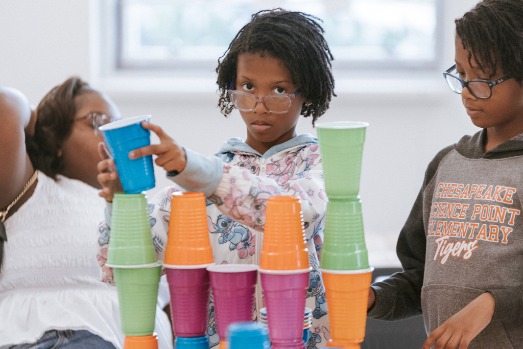 AACPS Mentoring Conference 2026: wo children stack colorful cups while another watches, all wearing glasses. The background features a blurred person.