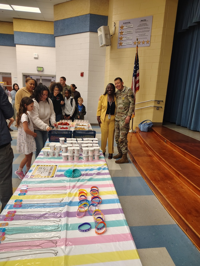 Indoor area with tables. A uniformed person stands next to a woman and child. Items on a table include bracelets and cups.