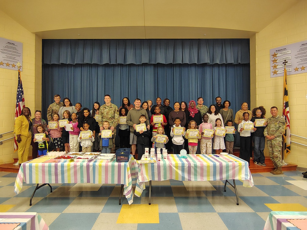 Group of adults and children holding certificates, standing together in a room with tables, stage, and flags.