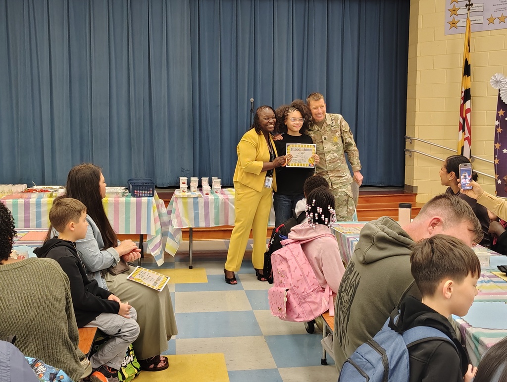 Group of people in a room with tables and chairs. A woman in yellow gives a certificate to a woman in a military uniform.