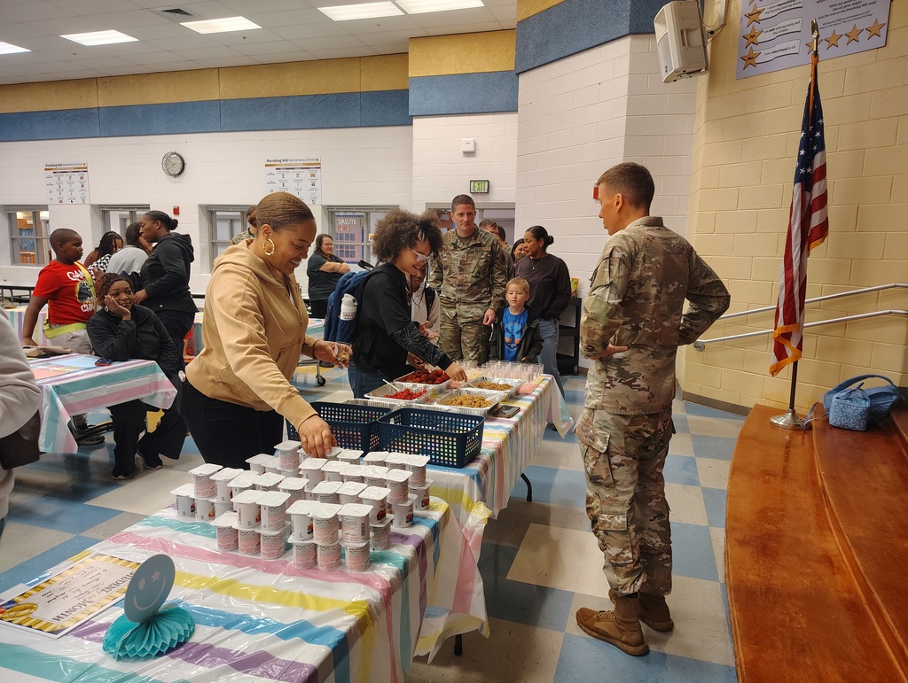 A military member in uniform speaks with a woman in a beige jacket at a table with food.