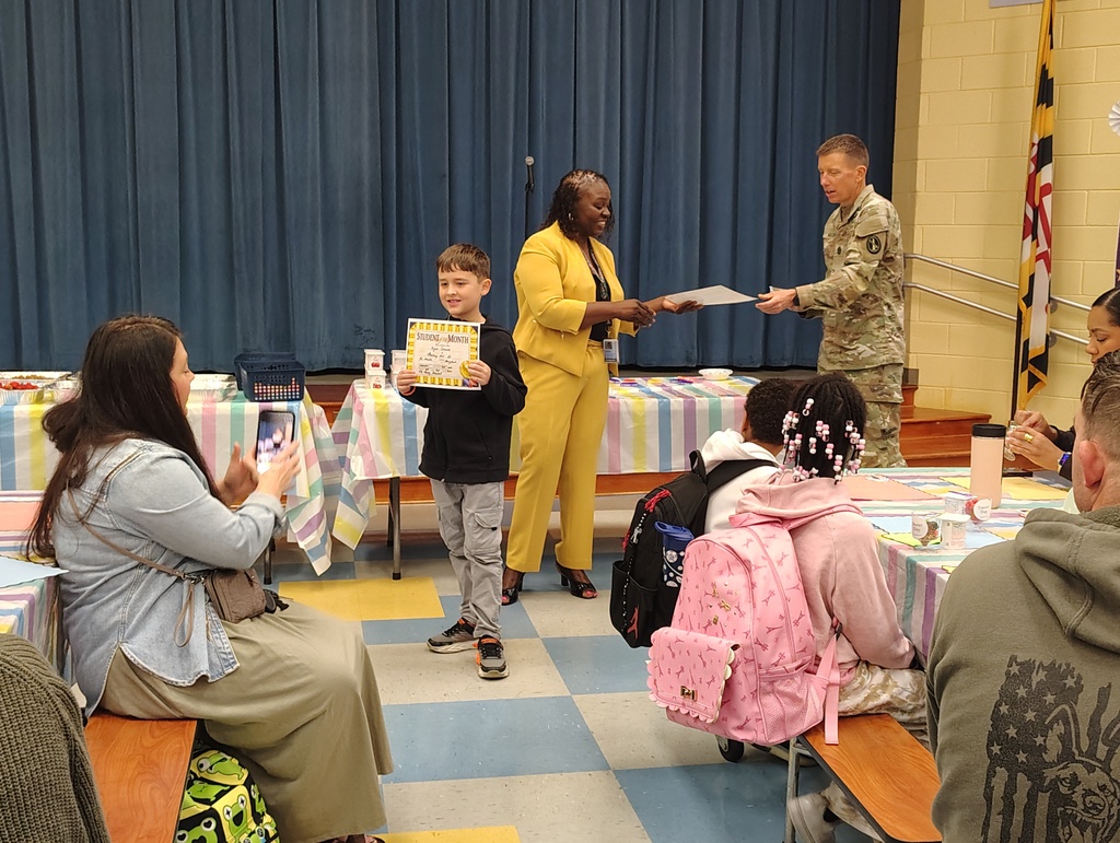 People seated at tables in a room, a man in uniform hands a certificate to a boy, others watch.