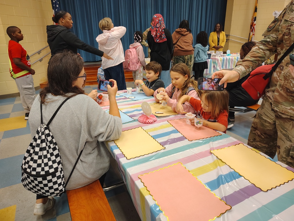 A group of people are seated at a table with food, while others stand and move around in a room.