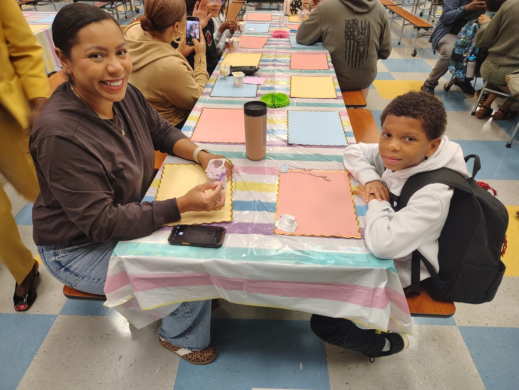 A woman and a child sit at a table with various colored papers, a cup, and a backpack.