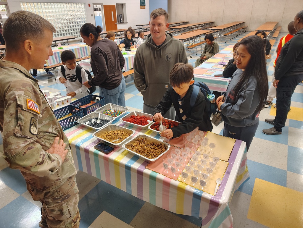 A military person stands among students. A child eats from a tray. Another child holds a backpack.