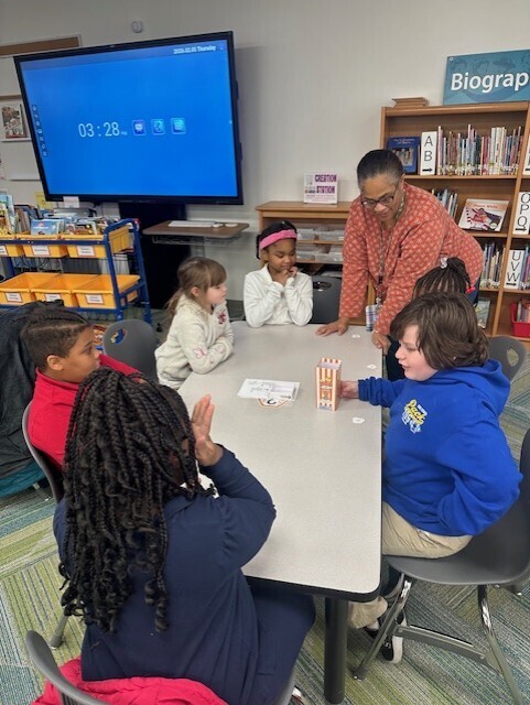 A classroom with children sitting around a table. A teacher stands behind them. A monitor is on the wall.