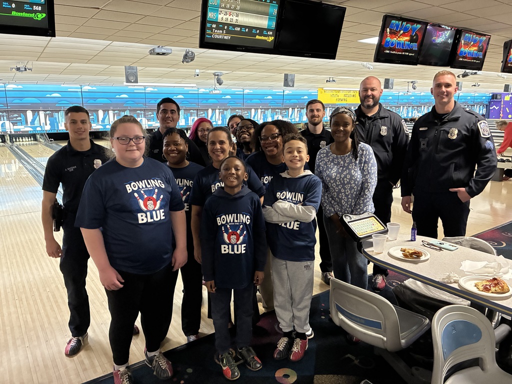 A group of people in matching blue shirts stand together in a bowling alley, with two adults in uniforms.