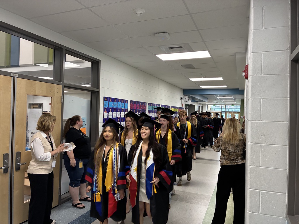 Graduates in caps and gowns walking in a hallway with some standing in doorways and others observing.
