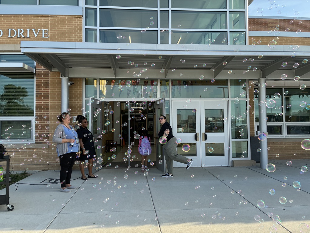 People stand outside a building with a canopy and bubbles in the air. The building has brick walls and glass windows.