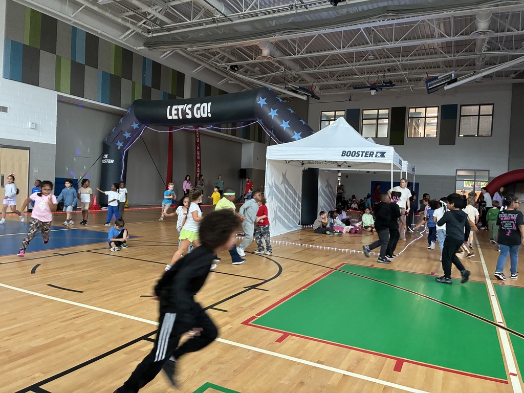 A group of children are playing in a gymnasium. Some run on the floor while others sit on the sidelines.