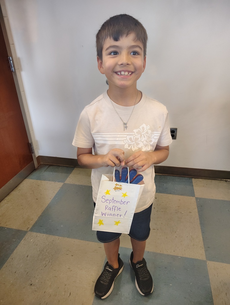 A smiling child in a white t-shirt, shorts, and sneakers holds a bag with "September" written on it. Behind, a white wall and an open door.