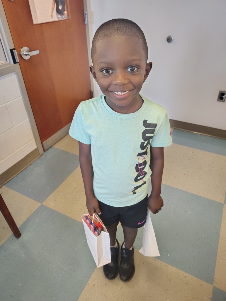 A child wearing a light blue shirt, black shorts, and black shoes holds two bags in a hallway with a checkered floor.