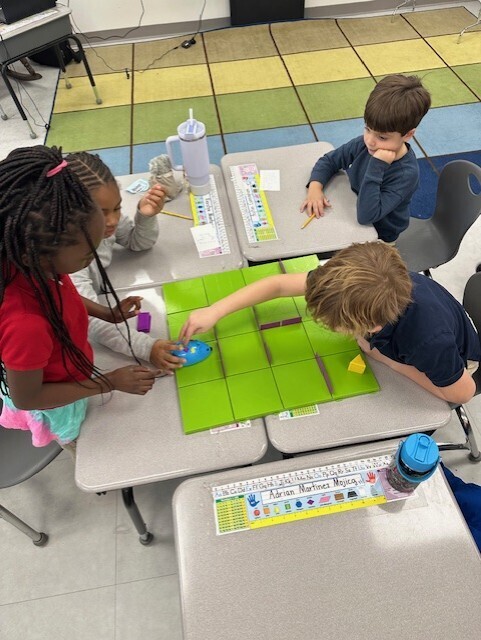 Four children seated at a table, engaged in an activity involving tiles, a board, and a small object.