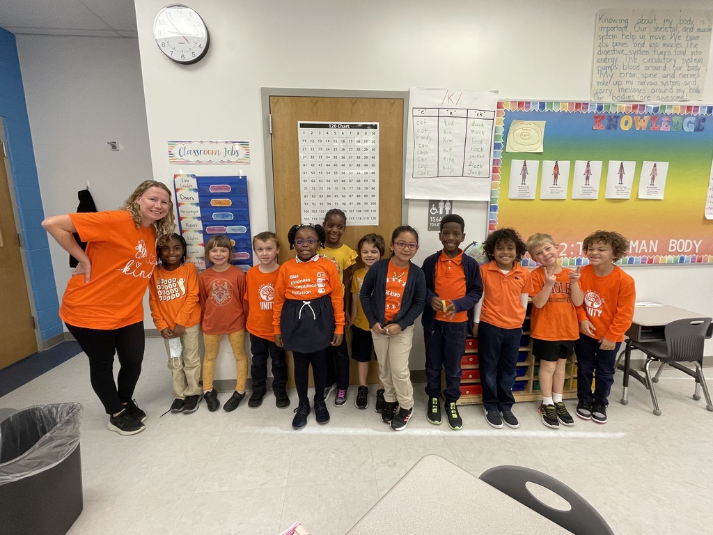 A group of children in orange shirts pose in a classroom, with a teacher standing to the left.