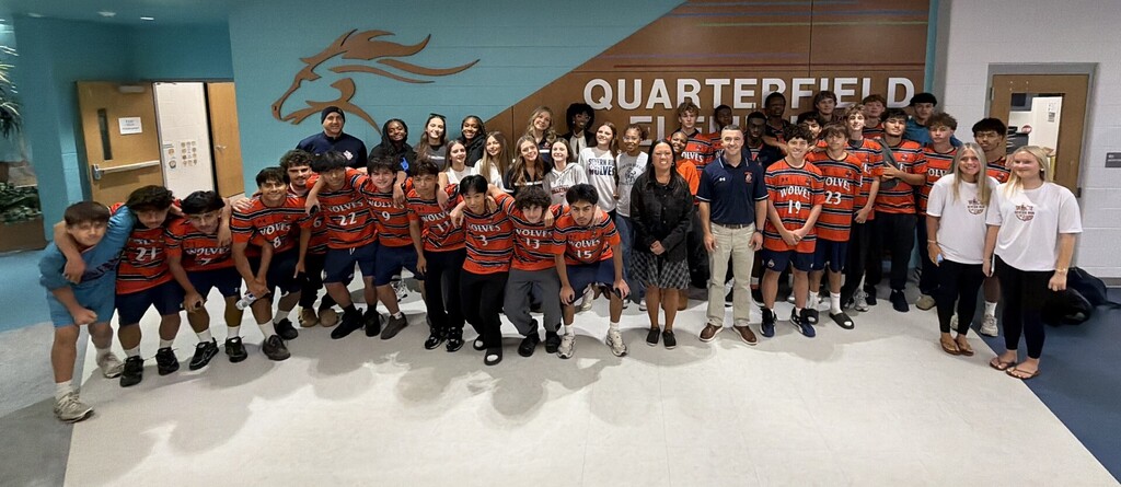 A team wearing orange jerseys poses for a photo with two adults in front of a wall.