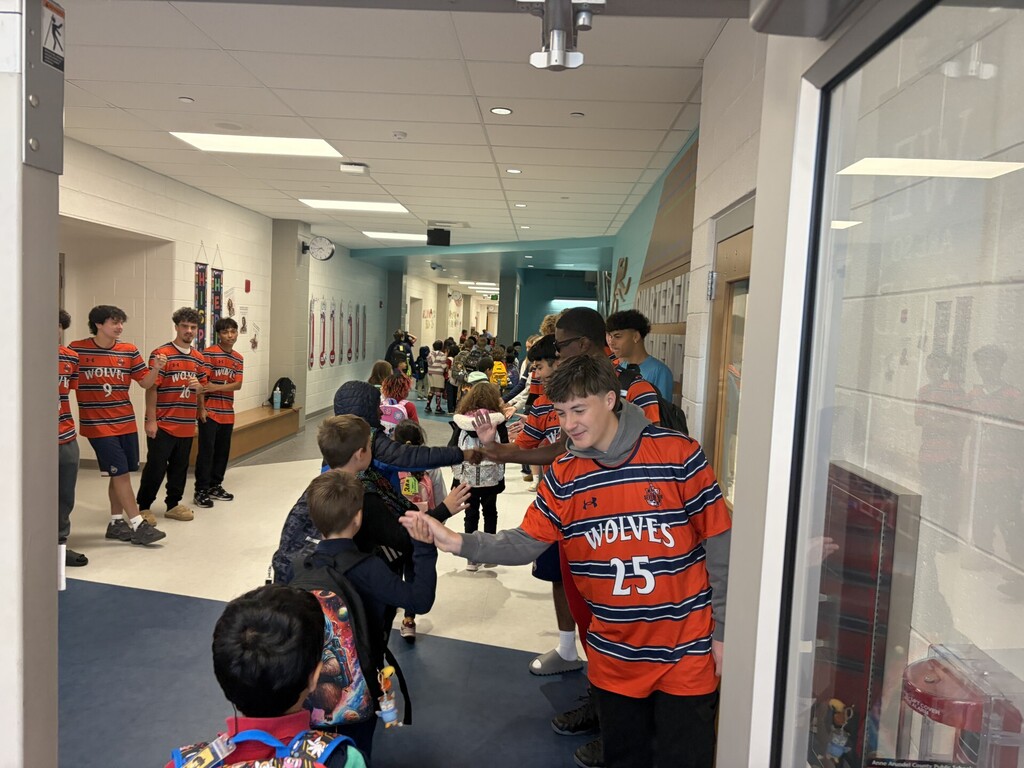 A group of kids wearing orange and black shirts gather in a school hallway. One child shakes hands.