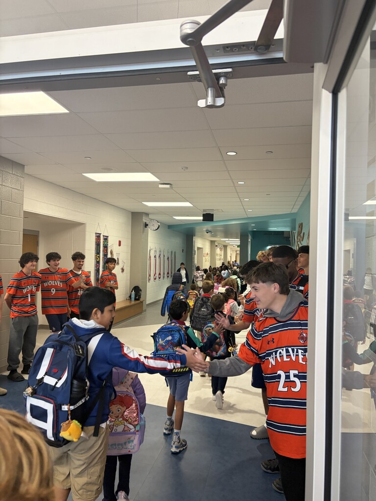 Students wearing uniforms and carrying backpacks are in a school hallway, greeting and shaking hands.