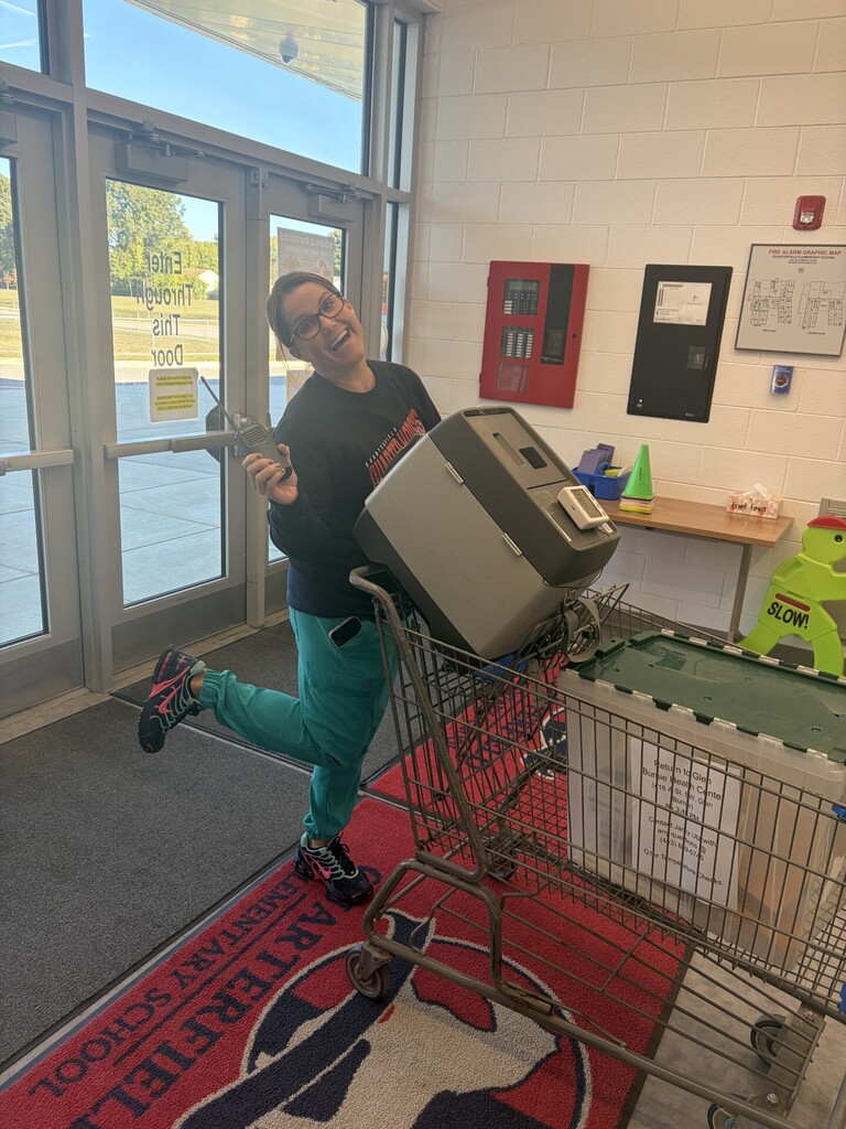 Person pushing a shopping cart with a printer on top, wearing a black shirt and green pants.