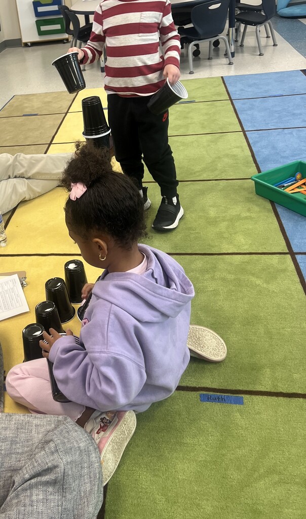 Two children in a classroom stacking cups on a carpeted floor, one wearing a hoodie.
