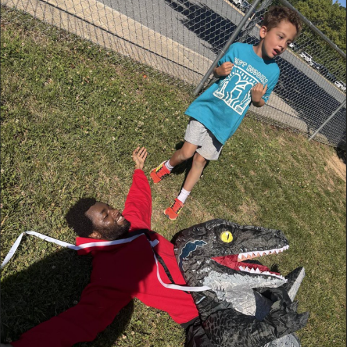 A student stands over a staff member dressed in an inflatable dinosaur costume who is exhausted lying on the ground after competing in a race