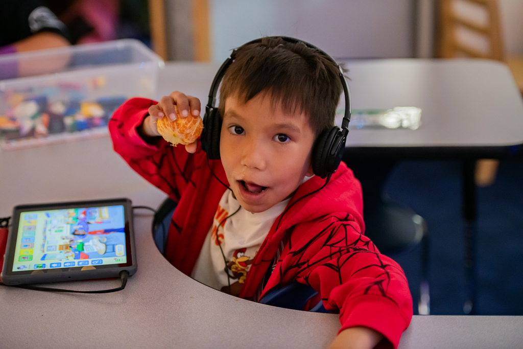 student eating a tangerine in a classroom.  he is wearing headphones and looking at an ipad