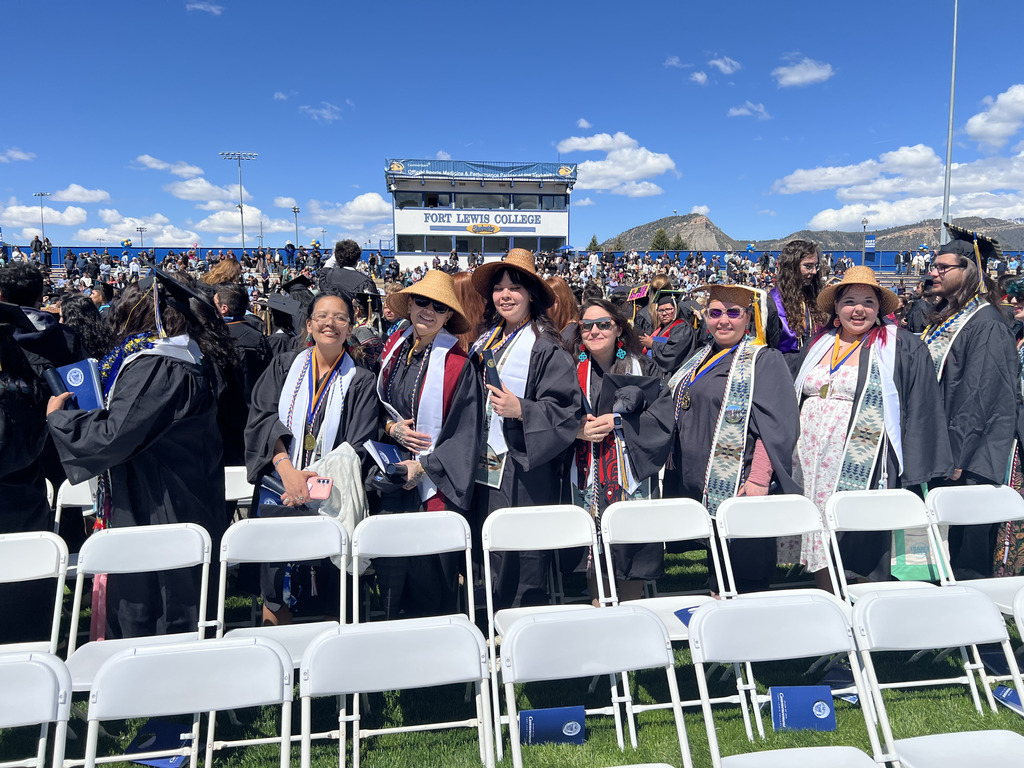 students in graduation caps and gowns, some wearing cedar hats, in the sunshine at a graduation ceremony