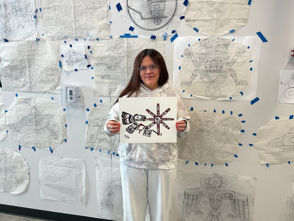 student holding formline painting in front of wall of formline drawings