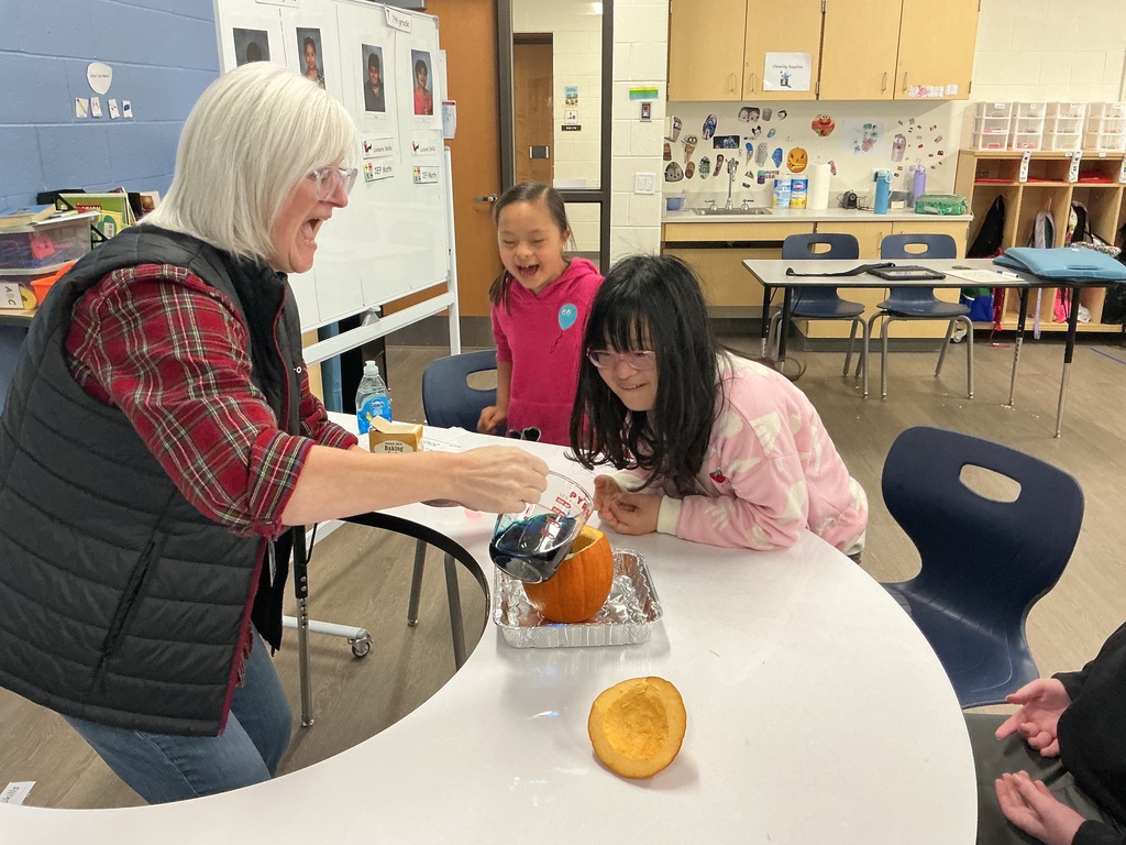 Little girls watching the teacher pour a liquid into a pumpkin.