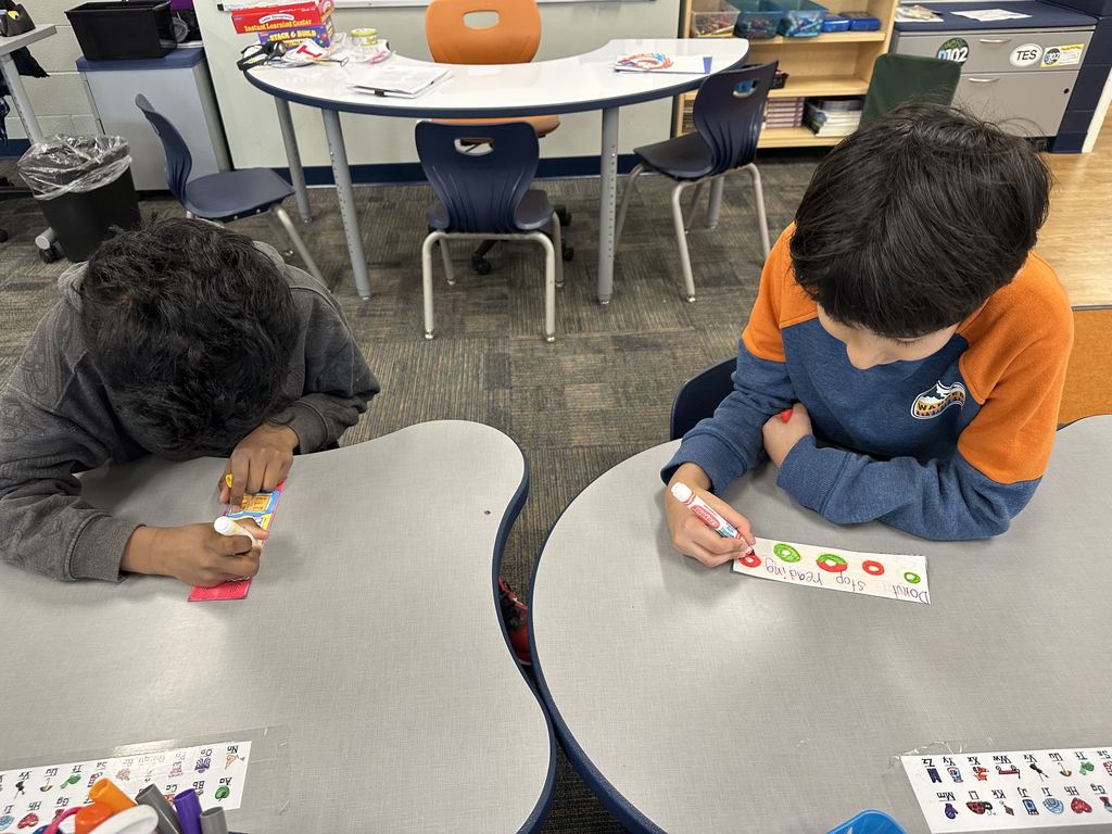 Two boys sitting at a desk and coloring bookmarks with markers.