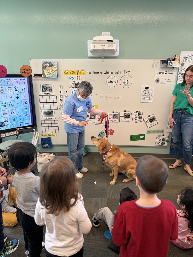 Dog owner stands with the dog, encouraging it to do a trick. Students are watching.